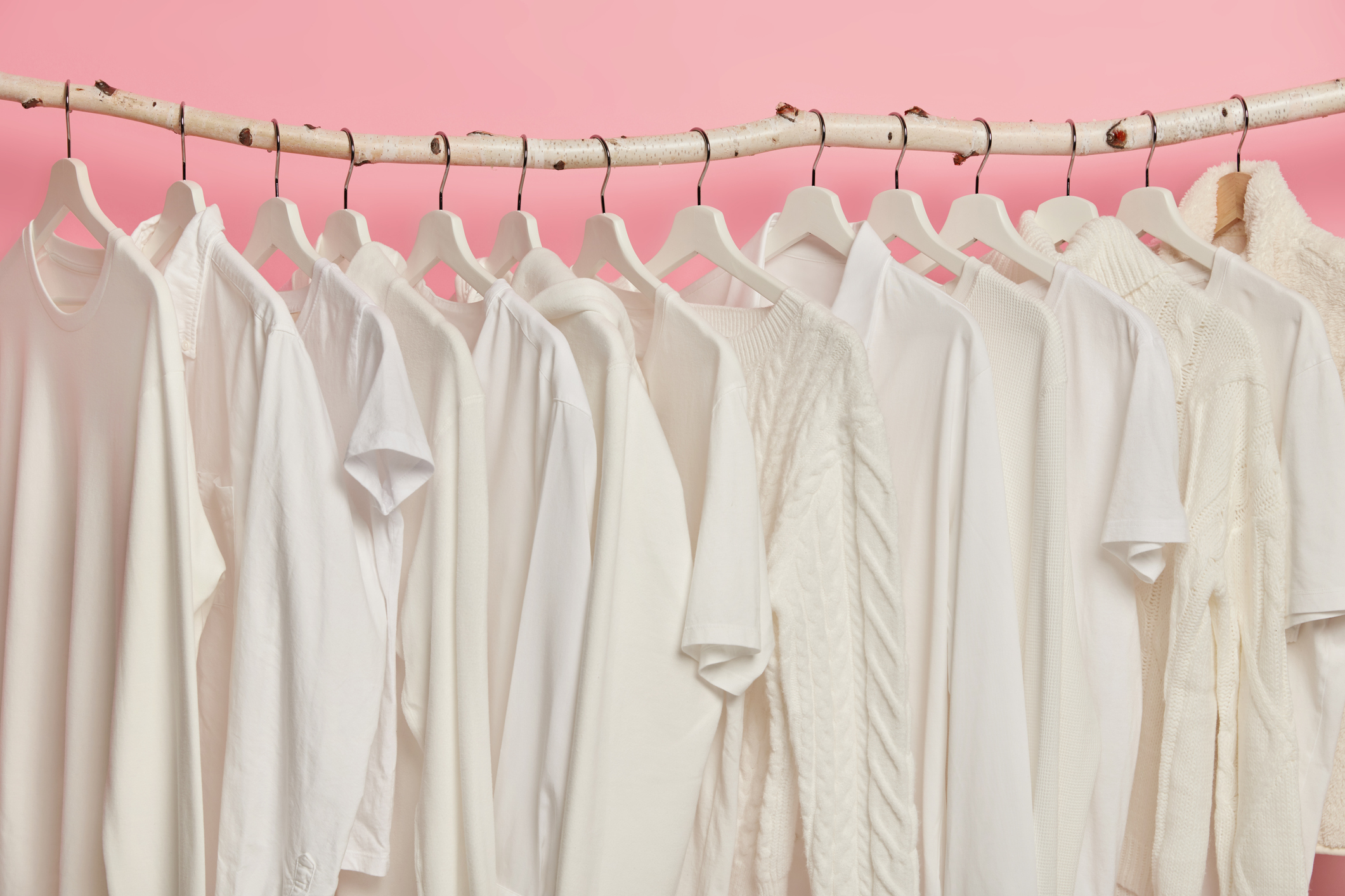 White solid clothes hanging in one row on wooden racks against pink background.